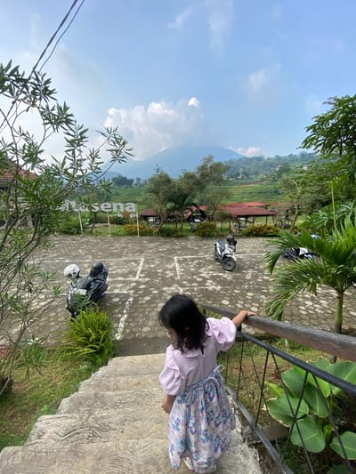 A young girl with dark hair, wearing a light pink shirt and a light blue floral skirt, stands on a concrete platform and leans against a metal railing, looking down. The location appears to be a scenic overlook or viewing area in Penanggungan, Indonesia. The ground is paved with hexagonal concrete blocks, marked with white painted parking spaces. Several parked motorcycles are visible in the parking area. Beyond the parking area, there is a view of rice paddies, trees, and mountains. There are also small structures with red roofs visible amongst the trees. The sky is blue with scattered clouds. Power lines are visible in the foreground. Vegetation grows along the edge of the platform where the girl is standing.