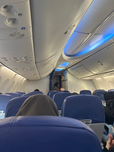 The image shows the interior of an airplane cabin. The viewpoint is from a passenger's seat, looking forward. Rows of blue airplane seats are visible, some occupied by passengers whose heads and shoulders are seen. Above the seats, the overhead compartments and ceiling panels are visible. There is blue LED lighting along the upper curvature of the cabin. A flight attendant or passenger can be seen standing near the front of the cabin, in the aisle. Safety instructions and signs are affixed to the panels above the seats. The scene is indoors, within a mode of transportation. The location is identified as Baji Mangngai, Indonesia, which is a village in South Sulawesi, Indonesia.