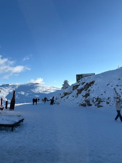 This image depicts a snow-covered winter scene on Uludağ Mountain, located in Bursa, Turkey. The foreground and midground are covered in deep white snow, forming a wide, open area or path. The sky is bright blue with minimal scattered white clouds.

Approximately 10-15 individuals are present, most of whom are walking on the snow-covered ground. Their attire consists of winter clothing, predominantly dark, though specific details are indistinct due to distance. One individual in the far right foreground is walking from right to left, wearing a light-colored jacket and dark pants, and appears to be speaking on a mobile phone. A small cluster of people is gathered in the midground, slightly right of center, with one person stooping or kneeling in the snow.

On the left side of the foreground, a tall, dark, bottle-shaped sculpture stands upright in the snow, beside which a red pole is partially visible. A dark wooden bench, partially snow-covered, is positioned in the immediate left foreground. To the right, a rocky hillside covered in snow rises, featuring a small, rectangular stone structure with a dark roof built into its slope. A single snow-laden conifer tree stands near this structure. In the distant background, snow-covered mountains exhibit structures suggestive of a ski lift or cable car system. The overall environment indicates a sunny, cold winter day in a mountain resort setting.