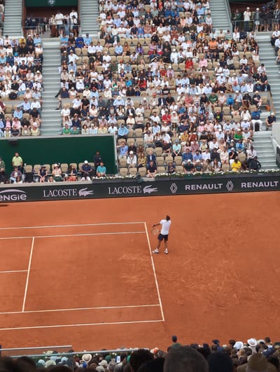 The image depicts a tennis match taking place on a red clay court. A male tennis player in a white shirt and dark shorts is positioned on the court, with his racket in hand. The court is lined with white markings. A crowd of spectators is seated in the stands surrounding the court. Advertisements for Renault, Lacoste, and ENGIE are displayed along the perimeter of the court. The scene is likely taking place in Boulogne-Billancourt, France, given the presence of the Roland Garros stadium, a well-known tennis venue in that location.