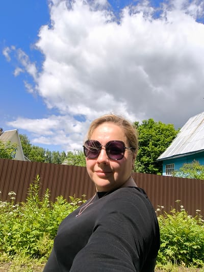 In a medium shot, a woman with blonde hair and sunglasses is seen in an outdoor setting. She is wearing a black top with a necklace. Behind her, a brown corrugated metal fence runs horizontally across the frame. Lush green foliage with small white flowers grows in front of the fence. In the background, trees, a house with a white roof and blue siding, and another house with a light-colored roof are visible. The sky above is blue with large, white cumulus clouds. The scene appears to be in the village of Novonikolskoye, Russia, likely during the daytime.