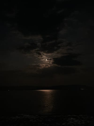 The image captures a nighttime scene at Ofrynio Beach, Greece, under a moonlit sky. The moon is partially obscured by clouds, casting a soft glow on the water and creating a shimmering reflection. The sea is calm, with a faint outline of the coastline visible in the distance, dotted with distant lights. The foreground is dark, hinting at the edge of the beach or a pier. The overall atmosphere is serene and tranquil.