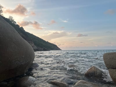 The image depicts a rocky beach scene near Baan Chaloklum, Thailand, at what appears to be either sunrise or sunset due to the soft, pastel colors of the sky and water. A large, rounded boulder is prominently located in the left foreground, partially submerged in the water. Other rocks, of various sizes, are visible near the waterline and in the water. The water is choppy with small waves breaking gently over the rocks. On the left, a tree covered hill or small mountain extends into the sea. The sky contains wispy, pastel-colored clouds.