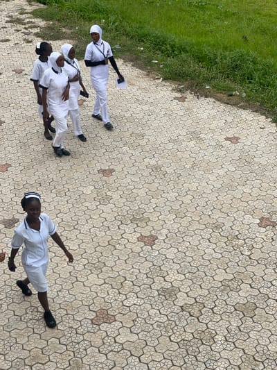 In Tavo, Nigeria, a group of five young women, presumably students, walk across a patterned concrete walkway. Four of the women, dressed in white uniforms and headscarves, are walking in a line together, some carrying notepads. The fifth woman, in a nurse's uniform with a striped cap, walks ahead of them, looking towards the camera. The walkway is bordered by a strip of grass and vegetation, with some scattered debris visible along the edge.