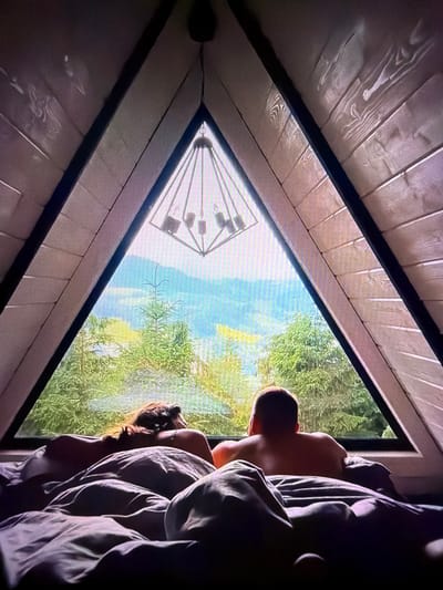 The image depicts an indoor scene inside a triangular-shaped structure, possibly a modern cabin or a unique architectural dwelling, overlooking a natural landscape. The foreground is dominated by a bed with dark gray bedding, where two individuals are lying down. On the left, a person with long, reddish-brown hair is resting their head on their arm, facing away from the camera and towards the window. To the right, another person, with short hair and a bare back, is also facing the window. Both appear to be relaxed and observing the view.

The focal point of the scene is a large triangular window that offers a panoramic vista of a lush green forest and distant mountains. The trees are dense and varied, with some appearing to be coniferous. The sky is overcast, casting a soft light on the landscape. Above the window, a modern geometric chandelier with multiple light fixtures hangs from the wooden ceiling. The interior walls are constructed from light-colored wooden planks, giving the space a rustic yet contemporary feel.

The scene suggests a peaceful and intimate moment shared by the two individuals as they appreciate the natural beauty from the comfort of their accommodation. The specific location within Beșalma, Moldova is not discernible from the image itself.