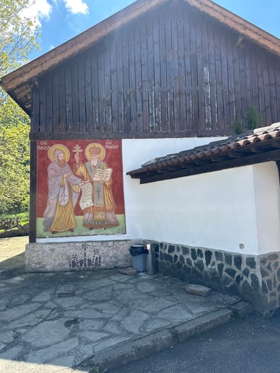 Here's a description of the image:

This is an eye-level, outdoor shot showing the exterior of a building. The building features a wooden gable roof on the left, adjacent to a white wall section. The white wall is adorned with a religious painting, featuring two figures with halos and robes, holding scrolls or books. A stone pathway is to the right of the painting and wall. The text "Стій КИРИЛЛ" and "Стій Медодій" in the top corner, suggests the subjects in the painting are saints. The scene seems to be taken in Barziya, Bulgaria. The image is rotated 90 degrees clockwise.