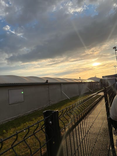 An outdoor scene at the Beauvais-Tillé Airport in Tillé, France, captures the late afternoon or early evening with a sunset casting golden light. A long, white tent structure with a curved roof runs horizontally across the middle ground. In the foreground, a black chain-link fence with vertical posts runs diagonally from the bottom right to the bottom left of the frame. Behind the fence, a strip of green grass is visible. The sky is filled with dramatic clouds, a mix of dark gray and lighter shades, with the bright sun peeking through the clouds on the right side of the frame, creating a lens flare effect. There are a few distant figures visible near the tent structure, suggesting activity. The overall impression is one of transition, possibly at the end of a day's events at the airport.