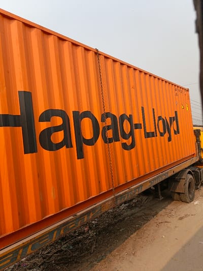 In Boliarpur, Bangladesh, an orange Hapag-Lloyd shipping container is depicted loaded onto a truck chassis. The container features vertical corrugations and displays the black "Hapag-Lloyd" logo prominently on its side. A metal chain hangs vertically along the container's side. Near the top right corner of the container, small white markings read "AV 1121" and "5.5," accompanied by a yellow triangular warning symbol. The truck's wheels and a portion of its yellow cab are visible on the right. The ground beneath the vehicle consists of uneven dirt and debris, with visible refuse or vegetation beneath the container on the left. The sky is a light, clear hue. No individuals or specific actions are observed in the scene.