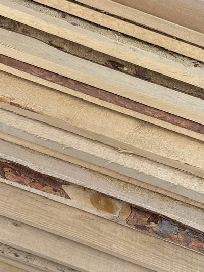This close-up, high-angle shot captures a dense arrangement of light-colored wooden planks or boards, stacked diagonally. The timber appears to be raw or roughly sawn, with varying textures; some planks are smooth, while others prominently display coarse saw marks and patches of reddish-brown bark along their edges. Abundant light-brown sawdust is visible in the gaps between the planks, suggesting recent cutting or processing.
The planks, likely pine or spruce given their color and the location context, are arranged in parallel lines, creating a repetitive pattern across the frame. The overall color palette is dominated by various shades of beige and light brown, with accents of darker brown from the bark. The lighting is soft and even, casting no distinct shadows, which might indicate an overcast day or diffused indoor light.
No human subjects, activity, or discernible events are depicted, as the image focuses solely on the stacked lumber and its textural details. There is no visible text. Given the location context of Kuda, Russia, the scene likely represents a stockpile of timber at a sawmill, lumberyard, or a construction site, emphasizing the raw materials of the region's industry.