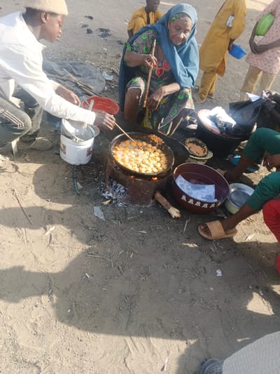 The image captures an outdoor food preparation scene in Gobirawa, Nigeria, under bright daylight. In the foreground, an elderly woman, dressed in a patterned garment and blue headscarf, squats on the ground, actively stirring spherical food items frying in a large metal pan filled with bubbling oil. The pan is positioned on an improvised metal drum stove, fueled by an active wood or charcoal fire. To her left, a young male in a white shirt, dark trousers, and a light-colored cap, kneels, holding a utensil over the pan and tending to a white plastic bucket. Scattered around the central activity on the unpaved, dusty ground are various containers including a red plastic bowl, a dark red patterned bowl, and a smaller metal bowl. In the background, three additional individuals are present: one seated wearing a yellow traditional garment, another standing in light brown attire holding a blue bowl, and a third with only their legs visible in green trousers and light brown sandals. The ground is dry earth with scattered debris and prominent shadows.