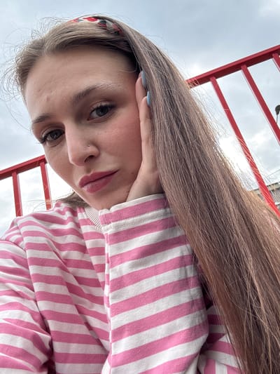 The image is a close-up, vertically oriented selfie of a young woman with long, brown hair. She is wearing a pink and white striped long-sleeved shirt. Her left hand is resting on her cheek, and she has a light blue nail polish on her fingernail. She has on eyeliner and mascara, and a small stud in her nose. The background shows a red metal structure, possibly part of an outdoor playground or park equipment, and a cloudy sky. The lighting suggests it is daytime, likely overcast. The image appears to be taken outdoors in Bylovo, Russia. The woman's expression is neutral. There is no visible text in the image.