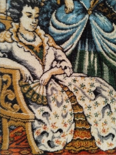 The image displays a close-up section of a textile, consistent with a rug or tapestry, depicting an artistic scene. A seated female figure, rendered in a style suggestive of 18th-century European portraiture, dominates the left and central frame. She is attired in an elaborate, high-necked cream-colored gown featuring ruffled sleeves and a full skirt decorated with repeating patterns of small orange and green floral motifs. A dark green panel with golden trim is visible on her chest. Her dark hair is styled upwards. Her right hand, positioned centrally, holds an open, segmented fan with green and white patterns. She is seated on an ornate chair with a golden-brown frame and scrolled armrests, part of which is visible on the lower left. The background on the upper right features a large, flowing drapery in shades of blue. Below this, behind the figure, is a dark green element with vertical striations or folds. The lower foreground beneath the figure's skirt shows an orange-red surface with subtle patterning. The scene depicted is an artistic rendering and does not show a specific real-world location within the city of Sucina, Spain.