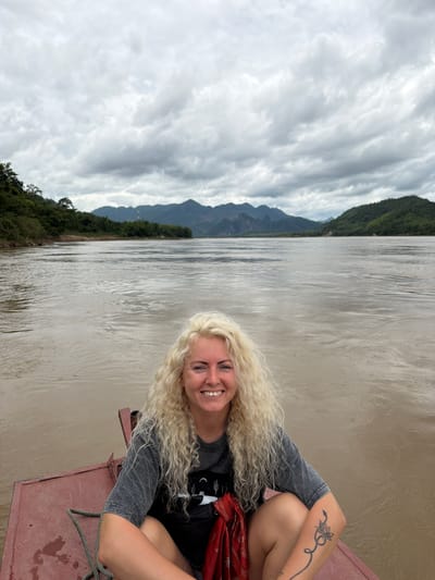 A woman with long, curly blonde hair is seated on the front of a red boat on a wide, muddy river. She is smiling and looking at the camera. She is wearing a grey t-shirt and black shorts. She has a tattoo of a snake and lotus flower on her right arm. In the background, there are green mountains and a cloudy sky. The scene appears to be in a rural area near Xanghay, Laos.