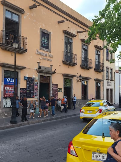 The image shows a street scene in the city of Querétaro, Mexico. The main architectural feature is a two-story peach-colored building with dark-framed windows, some of which have wrought-iron balconies. The building is identified as "Corregidora No.1 Centro Botanero" with the sign above the entrance. To the left of the building is a YALE store with a banner advertising sales.
On the sidewalk, there are pedestrians walking and standing. There are people of various ages and genders. The street is cobblestone. In the foreground, there are two yellow taxis with the license plates "A-882-TGH" and "A-644-TGJ". There is a woman walking in front of the taxis. A street lamp is near the YALE store, and another is seen further down the street. Trees are partially visible on the right side of the frame. The sky is partly cloudy.