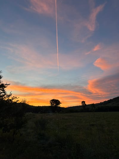The image shows a rural landscape at sunset near Debelets, Bulgaria. The sky is filled with vibrant colors of orange, pink, and blue, with wispy clouds. A contrail from an airplane stretches vertically across the sky. In the foreground, a dark, grassy field is visible, with silhouetted trees and bushes along the horizon. A gently sloping hill forms the background, also covered in dark vegetation. The overall scene conveys a sense of peacefulness and natural beauty.