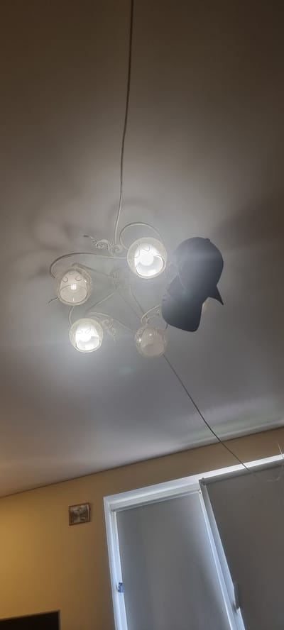 A black baseball cap is draped over a white, ornate ceiling light fixture with five glass globes. The cap hangs from the fixture, obscuring one of the lights. The light fixture is suspended from a thin cord that runs vertically along the ceiling. In the background, a wall painted a light yellow color is visible, with a small framed picture hanging on it. To the right, a window with a closed white blind is partially visible. The overall impression is an indoor residential setting.