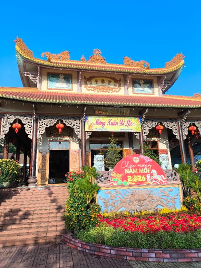 The image displays the exterior of Chùa Bảo Lâm (Bao Lam Pagoda) in Thành phố Tuy Hòa, Vietnam. The building exhibits traditional Vietnamese Buddhist architecture with a multi-tiered, ornate roof featuring red tiles and intricate gold decorations. The facade includes carved wooden elements, pillars, and two pictorial plaques depicting Buddhist figures on the upper level.
A yellow banner across the building's front displays "CHÙA BẢO LÂM," with another banner below it reading "Mừng Xuân Di Lặc." Red lanterns are suspended near the main entrance. A wide set of red-brown brick steps leads up to the pagoda's entrance. The area surrounding the steps and the building's base is adorned with various potted plants, including yellow chrysanthemums and kumquat trees, along with planted red and yellow flowers. Two white, stylized lion statues flank the central entrance.
In the foreground, a prominent New Year display is situated. It consists of a large blue relief depicting a crab-like motif, topped by a semi-circular red sign. This sign is inscribed with "Chúc mừng NĂM MỚI 2026" (Happy New Year 2026) and features a graphic of a white horse. The scene is illuminated by natural light under a clear blue sky. No individuals are visible in the image.