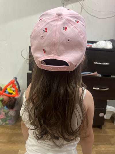 The image shows the back of a young child wearing a pink baseball cap with embroidered cherries. The child has long, brown hair that is visible from under the cap and falls down their back. They are wearing a white t-shirt. The child appears to be indoors, with a dark dresser with shiny handles in the background. A white object, possibly a changing table or a stool, is also visible in the background, along with a colorful toy in the lower right corner of the image. The scene is dimly lit, suggesting it might be daytime or evening. The overall impression is a casual, domestic setting, likely a home. No text is visible in the image.