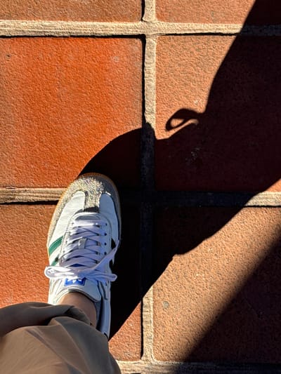 An overhead shot on terracotta paving stones. A white shoe with green stripes and white laces is partly visible, encroached upon by a large, dark shadow with an odd shape near the corner. The shadow extends over three paving stones. Beige pants are visible along the right edge, indicating the wearer's presence. Bright sunlight creates sharp contrasts, highlighting the texture of the bricks and the shoe.