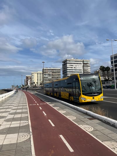 The image captures a bright yellow bus on a city street in Las Palmas, Spain. The bus is positioned to the right of a red bike lane, separated by a low concrete barrier with a semicircular design. The license plate is visible, but the details are difficult to discern.
Behind the bus, modern multi-story buildings rise into a sky dotted with clouds. Street lights line the street, and in the distance, people are visible along the pathway. The overall scene suggests a coastal urban environment on a clear, sunny day.