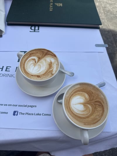 Two cups of latte art coffee are placed on a white tablecloth at a table. In the background, a green menu with gold lettering is visible. The text on the menu reads "THE PLAZA" and a logo. A white paper menu with "THE I DRINK." is under the coffee cups. The bottom part of the paper menu shows social media information with a Facebook logo and text that reads "The Plaza Lake Como." This scene is likely located at The Plaza Hotel in the city of Como, Italy.