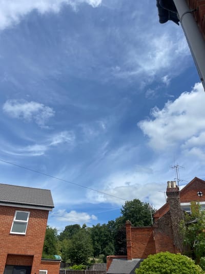 A low-angle, wide shot shows a bright blue sky with wispy white clouds. In the foreground, the roofline of two red-brick houses is visible, one on the left with a gray tiled roof and a white-framed window, and another on the right with a chimney and an aerial. Power lines stretch across the sky, dividing it horizontally. Lush green trees form a backdrop behind the houses. The scene appears to be in a residential area.