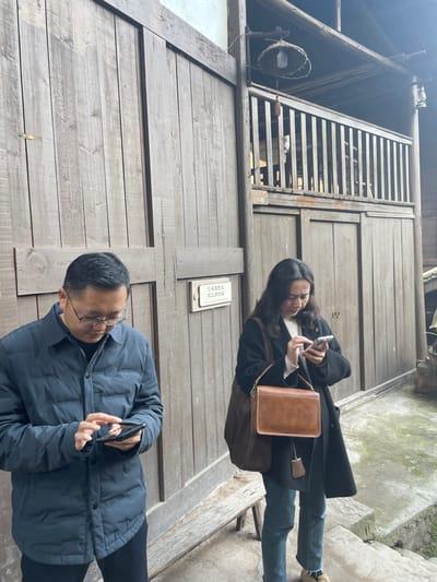 Two individuals stand in front of a wooden structure. The person above, wearing a black coat and jeans, is holding a cell phone and carrying a brown bag. The person below, dressed in a blue jacket and black pants, is also holding a cell phone. The wooden structure includes a staircase and an enclosed balcony. A sign is visible on the wall. The ground appears to be a combination of concrete and stone.