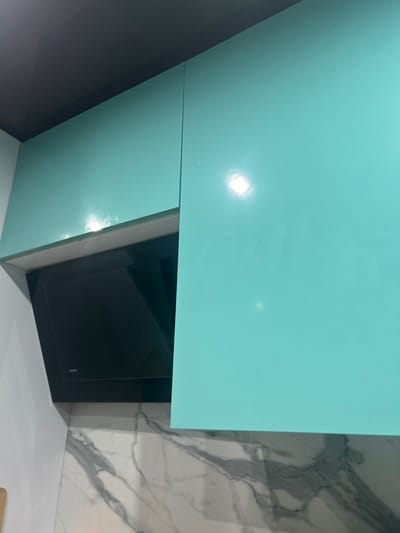 Dmitriyevskaya, Russia. An interior scene, likely a kitchen, is depicted with a vertical orientation. The upper portion of the image shows two glossy turquoise upper kitchen cabinet doors. The left cabinet door is partially open, revealing a dark void behind it, while the right cabinet door is closed. Bright reflections of light are visible on the polished surfaces of the cabinets. Beneath the left cabinet and partially extending into the center is a black, slanted, glass-front range hood, with the brand name "CATA" visible in small white text on its lower left side. A white wall is visible to the immediate left of the range hood. Below the cabinets and range hood, a countertop or backsplash with a white and grey marble pattern is present. The ceiling at the top of the image is dark grey. No individuals are present in the scene.
