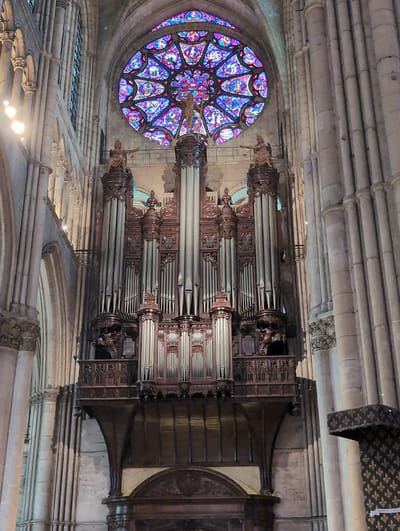 This image shows a large pipe organ located inside a cathedral. The organ is ornate, made of dark wood and polished metal pipes. Above the organ is a large, colorful stained-glass rose window, which illuminates the interior with vibrant blues, purples, and reds. The cathedral walls are constructed of large stone pillars and arches, indicative of Gothic architecture. Statues of figures are positioned on top of the organ's framework. The lighting is dim, with some artificial lights visible, contributing to the solemn atmosphere of the religious building. There are no people clearly visible in the image. The location is the Church of Saint-Nicolas in Urvillers, France.