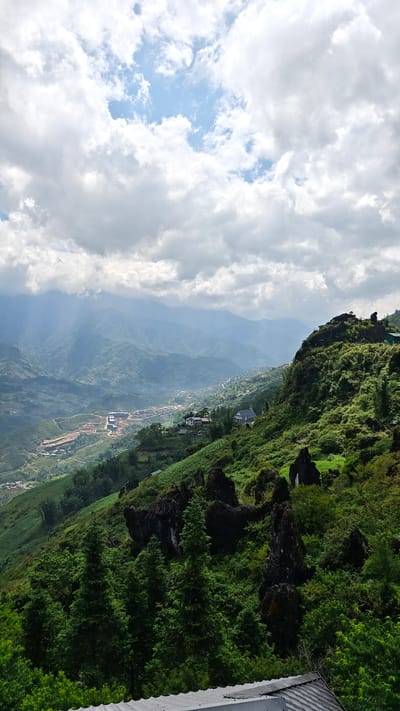 A landscape view in Tả Van, Vietnam shows a lush, green hillside in the foreground. A variety of trees and vegetation cover the slope, interspersed with dark, rocky outcrops. In the distance, a valley is visible with a small settlement and terraced fields. Further away, a mountain range fades into the hazy background. The sky above is filled with a mix of heavy, grey clouds and patches of blue sky. The overall light is diffused, suggesting an overcast day.