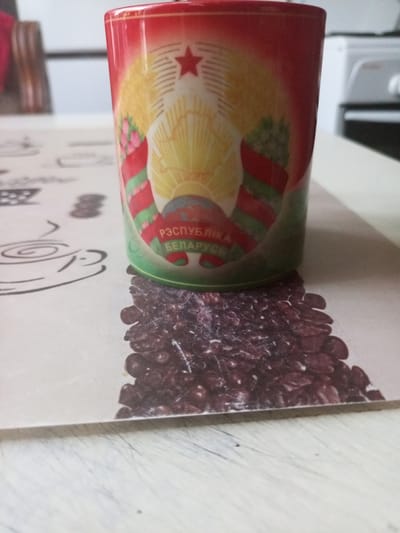 A close-up, eye-level shot features a cylindrical mug adorned with the coat of arms of Belarus, positioned on a table with a patterned surface depicting coffee beans and coffee cup outlines. The mug's design includes a red star at the top, a sunburst, and a ribbon with the inscription "РЭСПУБЛІКА БЕЛАРУСЬ" (Republic of Belarus). The mug has a gradient of red at the top transitioning to green at the bottom. The table surface beneath the mug is a beige-colored mat with a detailed print of dark red coffee beans. To the left of the mug, on the same mat, are faint black line drawings of coffee cups. The background is blurred, showing indistinct shapes of furniture and appliances, suggesting an indoor setting in a home or cafe. The overall lighting is moderate. The image captures the mug and its immediate surroundings on the table.