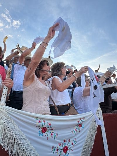 A crowd of spectators in San José del Valle, Spain, is waving white handkerchiefs in the air. The scene is outdoors, likely at a bullring or similar venue. The sky is visible, with a few scattered clouds. A white fabric barrier with floral embroidery hangs in the foreground. The crowd consists of both men and women of varying ages, many wearing hats and casual clothing. Some are wearing bracelets. The handkerchiefs are held up in apparent appreciation or support. One person has a white handkerchief with black writing on it. A few are standing behind a metal fence.