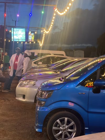 The image depicts an outdoor evening or nighttime scene, possibly at a roadside stop or market in Kimorori, Kenya. Several vans or minibusses are parked in a row, facing the viewer's right. The closest vehicle is a vibrant blue van, followed by lighter-colored (white or silver) vans. All the vehicles have decorative LED strip lights running along their sides, reflecting the ambient light.
In the foreground, two men are visible, both wearing white long-sleeved shirts or coats and white head coverings, suggesting uniforms perhaps for food service or transport. One man, closer to the center, wears a red identification tag. Another person's legs are partially visible further down.
The sky in the background is dark, indicating it's late evening or night. A string of warm yellow-orange bulb lights curves across the upper left portion of the frame, providing illumination. Other colorful individual lights (red, green, blue) are also scattered. In the mid-ground, behind the people, a brightly lit screen or sign is visible, displaying an image and some text. Above this, bottles with faint labels are placed on a shelf.
Visible text includes "JUAN" on the windshield of the blue van, with other letters possibly following (e.g., "MALA"). On the background screen, the text "TREAT WITH LIGHT" is legible. The ground appears to be unpaved, consistent with a rustic or informal setting. The vehicles resemble models commonly used for public transport in Kenya, known as matatus.