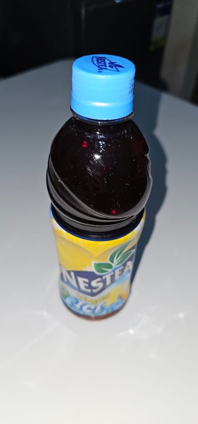 This is a close-up, low-angle, indoor shot of a bottle of Nestea Iced Tea, specifically the Lemon flavor, placed on a light-colored surface. The bottle is oriented horizontally, with the cap on the left and the body extending to the right. The cap is bright blue and features the distinct Nestea logo with two stylized leaves and the brand name "NESTEΛ". The bottle itself is dark brown, filled with the iced tea, and reflects light from the surroundings, showing some scratches and smudges on its surface.

The label on the bottle is predominantly yellow with blue and green accents. The "NESTEΛ" logo is prominently displayed on the label in blue and green. Partial text is visible on the label, including "LEMON" and parts of the word "ICE" in blue lettering, confirming the flavor. The lighting suggests it's not broad daylight, possibly late afternoon or evening, with the light source creating a shadow to the left of the bottle. The background is out of focus, consisting of dark and light areas, suggesting an indoor setting. There are no people visible, and the focus is solely on the product. The image quality is somewhat grainy, with red specks of light visible on the bottle, likely reflections or artifacts. The overall impression is a product shot, possibly for e-commerce or personal documentation, taken in a neutral, indoor environment in Makati, Philippines.