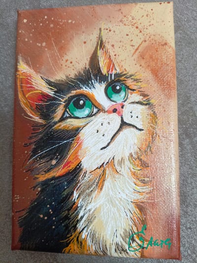 This is a close-up, vertical, eye-level shot of an acrylic painting on canvas featuring a calico cat's face. The cat is looking upwards and to the right with large, bright green eyes and a pink nose. The painting's background is a warm, mottled brown and tan with flecks of white and rust-colored paint splatters. The cat's fur is depicted with thick, textured brushstrokes in black, white, orange, and hints of yellow and red. Whiskers are rendered with thin white lines. The overall impression is one of curiosity and perhaps playfulness. In the bottom right corner of the painting, a green artist's signature is visible, appearing to read "Aareg" or similar. The canvas is placed on a textured gray surface, possibly a rug or fabric. The lighting is neutral, and no specific time of day or weather conditions are indicated. The painting appears to be a standalone piece, not part of a larger scene. The location context provided is Azov, Russia, but there are no visual cues within the image to confirm this.