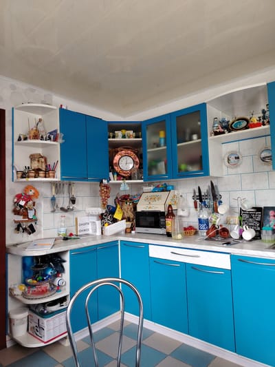 The image shows a cluttered kitchen in what appears to be an apartment in Rudzensk, Belarus. The kitchen features bright blue cabinets with white countertops and a checkered tiled floor. Various kitchen appliances and utensils are visible, including a microwave, a kettle, mugs on a drying rack, knives on a magnetic strip, and a large wall clock. Shelves are filled with decorative items, food containers, and bottles. A doll is hanging on the wall near a set of keys. The overall impression is one of a lived-in, functional, and somewhat disorganized kitchen space. There are no people present in the image.
