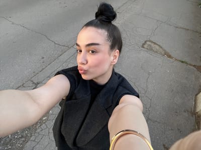 A person wearing a black sweater and a gold-colored bracelet is taking a selfie on a gray asphalt surface. The person has dark hair pulled back in a bun and is looking up at the camera with slightly pursed lips. The asphalt shows cracks and imperfections. The location is unknown, and no information is available in the image to determine if it is Niš, Serbia.