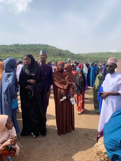 A medium-shot photograph taken outdoors shows a group of people standing or sitting on dirt ground in Naraguta Hausawa, Nigeria, during daylight hours. The scene is oriented vertically. A green, grassy hillside is in the background. A cloudy sky fills the upper portion of the frame.
Several individuals are visible, predominantly women wearing hijabs and other traditional Islamic attire. Colors range from brown and black to blue, orange, and white. A man in a white garment and cap stands at the edge of the group.
Some people are interacting while others are looking at the camera. No specific activities are readily apparent beyond the group gathering. A purse, a phone and keys are held.