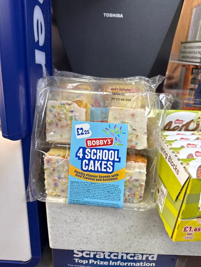 The image shows a package of "Bobby's 4 School Cakes" resting on a surface in what appears to be a retail setting in North Hertfordshire, United Kingdom. The cakes are vanilla flavored sponge cakes with iced toppings and sprinkles, packaged in a clear plastic container. A blue and white label on the package displays the product name, brand, price (£2.25), and ingredients. The cakes are likely for retail sale. The 'best before' date shown on the package is 18/06/26. Adjacent to the cakes are a box of "Sharing Blocks" and a scratchcard for Top Prize Information, also available in the area, suggesting the cakes may have been purchased at a retail location like a convenience store. A black "Toshiba" object and a blue "er" symbol are partially visible in the scene.