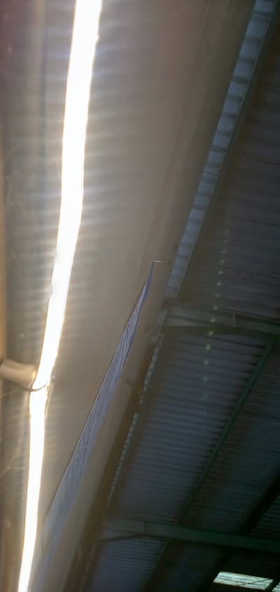 The image presents a perspective of a roof interior, likely of an industrial or commercial structure, possibly located in Banjaran, Indonesia, although location information isn't directly evident in the image. The roof construction features corrugated metal sheets supported by a framework of green-painted steel beams. A prominent, bright strip of light penetrates through a gap in the roof, casting strong shadows and highlighting the corrugated pattern. A narrow, possibly insulated panel runs along one side of the light gap. The angle suggests the photo was taken looking upwards towards the roof. No individuals, specific objects, or actions can be directly identified within the image besides the architectural elements.