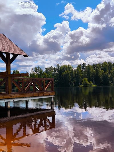An outdoor, vertical, eye-level shot captures a wooden pier extending into a dark lake on a cloudy day. The pier, made of light brown wood, has a slatted deck and a brown shingled roof structure on the left side. A wooden railing with diagonal supports lines the edge of the pier. The lake's surface reflects the sky, the trees on the far bank, and the pier itself. The far bank is lined with a dense band of green trees. The sky is a mix of bright blue patches and large, fluffy white and gray clouds. This scene is located at a lake in Vecumnieki, Latvia.