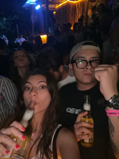 Two young adults pose for a selfie while drinking beer at night in what appears to be a crowded outdoor venue. A third person is partially visible in the background, also appearing to be drinking. The man wears glasses and a baseball cap, while the woman has long hair and red nail polish. They are holding Corona beer bottles. The environment is dimly lit with some lights visible in the background and the surroundings suggest that this venue is in Puerto Escondido, Mexico.