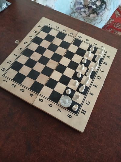 An overhead, eye-level, medium shot captures a wooden chessboard laid out on a dark brown, polished wooden table. The chessboard is divided into light and dark squares and is marked with letters A through H along one side and numbers 1 through 8 along the adjacent side. Several white chess pieces are positioned on the board, including pawns, rooks, and what appear to be bishops. One white disc-shaped piece is also present on the board. The background shows a portion of a patterned fabric, likely a pillow or throw, with floral motifs. The scene is located in Razzakov, Kyrgyzstan.