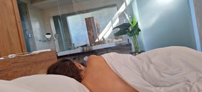 A person is lying in bed, facing away from the camera, covered by white bedding. The person's back and shoulder are visible. The scene appears to be a hotel room in Payangan, Indonesia, with a bathroom visible in the background. The bathroom features two sinks, mirrors, and a wooden cabinet. A large potted plant is also present. Sunlight streams into the room, creating shadows on the wall and floor. The overall impression is one of relaxation and a leisurely morning.