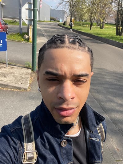A male individual is prominently featured in the foreground, looking directly at the camera. He has dark skin, short dark hair styled in cornrow braids, and a silver hoop earring in his left ear. He wears a dark blue denim jacket over a dark t-shirt, accessorized with a silver chain necklace. A light-colored strap with dark leather trim from a bag is visible over his right shoulder. The scene is set outdoors on an asphalt surface in Plaisir, France. To the subject's left, a blue and white sign is mounted on a green pole, with a red car parked behind it. Beyond the subject, an asphalt road extends uphill, bordered on the right by a grassy area with sparsely leafed trees and yellow flowers. Another person is visible in the mid-distance, walking away on the road. Industrial-style buildings with grey siding are present in the far background. The scene is brightly lit by natural sunlight under a clear sky.