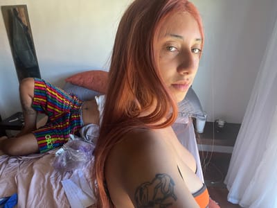 The scene depicts an indoor setting, identified as a bedroom in Arraial Do Cabo, Brazil. A female individual with long, light reddish-brown hair and dark eyes occupies the foreground, looking directly towards the viewer. She is wearing an orange and black bra top, and a tattoo depicting two interlocking figures is visible on her upper left arm.
In the background, a male individual is positioned on a bed with light pinkish-brown sheets and a grey duvet. He lies partially obscured, facing away from the camera, wearing multicolored striped shorts in yellow, green, pink, blue, and red, with the word "CHAOS" visible on the left thigh. A tattoo is present on his left calf. His upper body is largely hidden by a grey pillow and the hair of the female in the foreground.
On the bed, near the male's legs, a clear plastic bag containing purple material, possibly fabric or hair extensions, rests beside a crumpled piece of white paper. The room features white walls. To the left, a tall, narrow mirror with a wooden frame leans against the wall, draped with a dark jacket. To the right, a white translucent curtain indicates a window, admitting bright natural light. A dark-colored table or nightstand is visible beneath the window, holding a white cylindrical object, other indistinct items, and multiple wires.