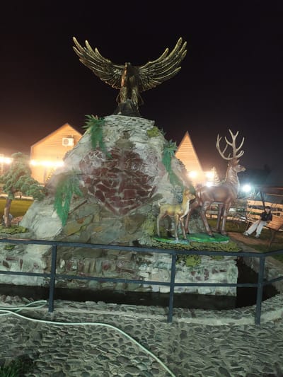 The image depicts a nighttime scene in Sosnovka, Kyrgyzstan, featuring an elaborate outdoor decorative fountain and landscaping. Dominating the central area is a large, multi-tiered rock structure, likely artificial, adorned with green, fern-like decorations. Atop this structure is a striking statue of a golden eagle with outstretched wings. At the base of the rock formation, two deer statues are positioned: a larger stag with an impressive rack of antlers and a smaller fawn.
The surrounding environment includes a building with a distinctive triangular roof, illuminated from within and by external lights, suggesting it could be a restaurant, cafe, or event space. To the left, a miniature, conical evergreen tree is visible. A dark, water-filled channel or moat runs in front of the rock structure, bordered by a black metal railing. The ground is paved with irregular stones, and a white hose lies on the paving stones.
To the right of the fountain, a wooden swing set is partially visible. A person, dressed in light-colored pants and a dark shirt, is seated on one of the swings, looking towards the left. The overall ambiance is enhanced by artificial lighting, creating a warm and inviting atmosphere despite the darkness of the night. The scene suggests a public recreational area or a hospitality venue.