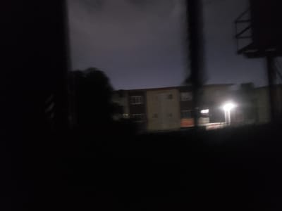 The image depicts a blurry, dimly lit urban scene at night. In the background, a multi-story building with visible windows and an orange-colored section can be seen. A bright light source illuminates a portion of the building and its surroundings. To the right of the building, a dark, angular structure resembling a billboard support is visible against the dusky sky. In the foreground and to the left, large, dark, out-of-focus shapes obscure the view, suggesting the presence of an object or structure closer to the camera. The overall impression is one of a nighttime street view in an urban area, with limited visibility due to darkness and motion blur. The location is identified as Iganmu, Nigeria.