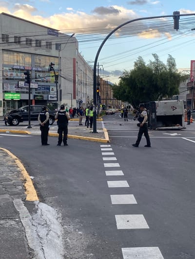 The image shows a street scene in Quito, Ecuador, with several police officers present. The time of day appears to be late afternoon or early evening, as indicated by the warm light and long shadows. The sky is partially cloudy, with some soft, golden clouds visible.
The main subjects are several police officers in uniform, some wearing vests with "POLICIA" clearly visible. They appear to be directing traffic or managing a scene, as some are standing in the road and others are observing. In the background, there is a large white truck, possibly a utility or delivery vehicle, and other vehicles on the street. Buildings line one side of the street, featuring various signs. One sign, partially visible, reads "farnacias Procómicas," and another indicates "DENTAL" services. The street is paved with asphalt and marked with white pedestrian crossing lines and yellow lines along the curb.
The overall impression is of an organized presence, possibly related to a traffic stop, an accident, or a public safety operation. The officers' postures suggest they are attentive and professional. There are no strong emotional cues visible, but the scene implies a regulated environment. The presence of multiple police officers and the truck in the road suggest an ongoing event that requires their attention.
