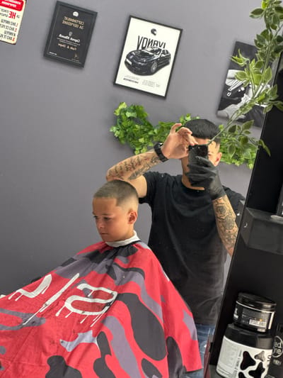 The media file is a vertical, first-person perspective photograph taken in a barber shop. The photographer, a man with tattoos on his arms and wearing a black t-shirt and black gloves, is using a phone to take a selfie in a mirror. He is in the process of giving a haircut to a young boy. The boy is wearing a red and black camouflage barber cape. In the background, the wall is adorned with framed pictures, including one of a car, and artificial green plants. On a shelf above, there are several hair product containers, one of which has a cow-print design and the text "One" and "MEN". The scene appears to be indoors, with artificial lighting. The overall mood is professional and focused on the barbering service. Visible text includes brand names and product descriptions on the hair products, as well as text on the framed pictures, some of which appear to be in Cyrillic script.